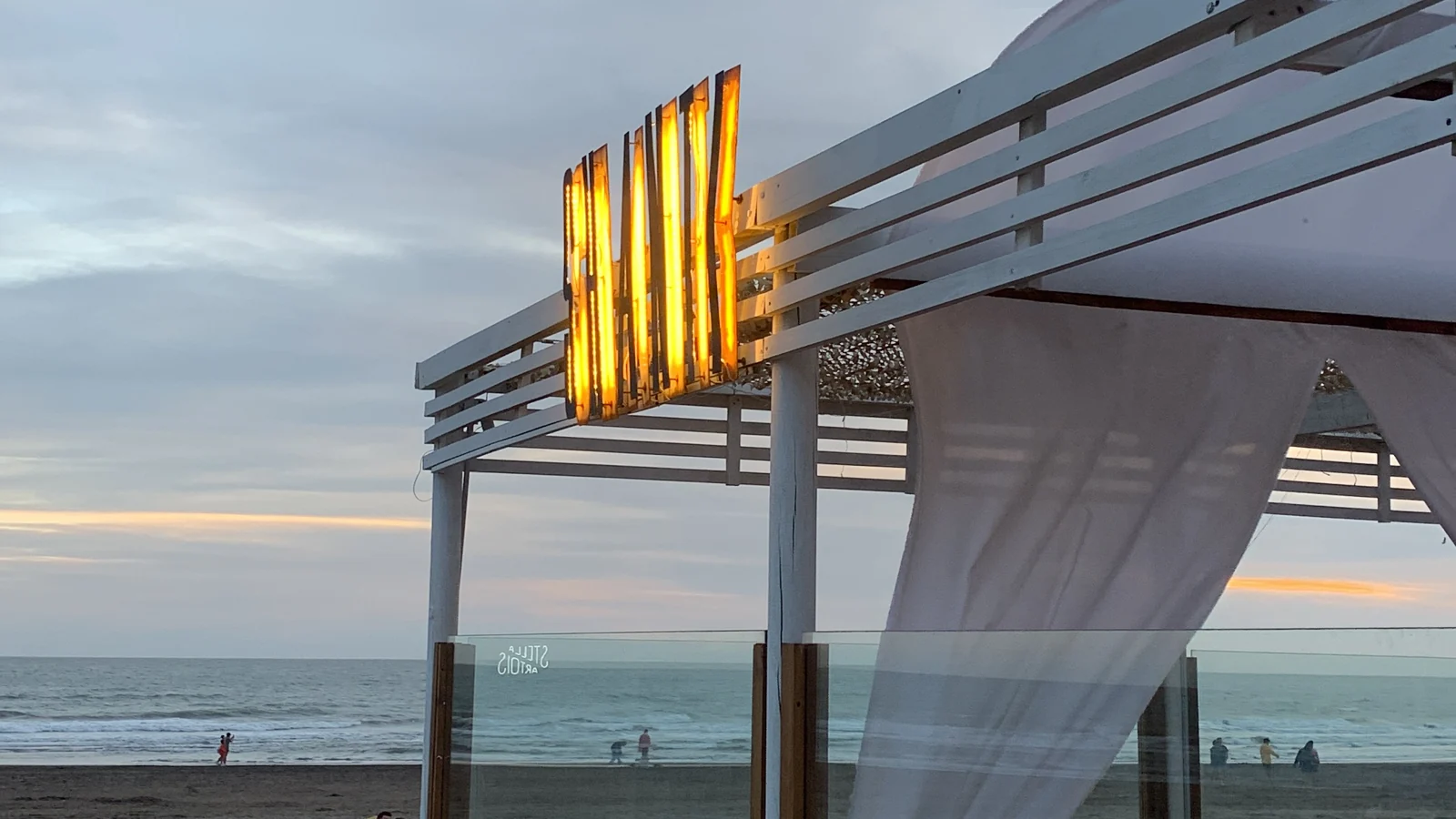 SHANTI al caer la tarde: luz cálida en la pérgola, telas blancas y mar al fondo.