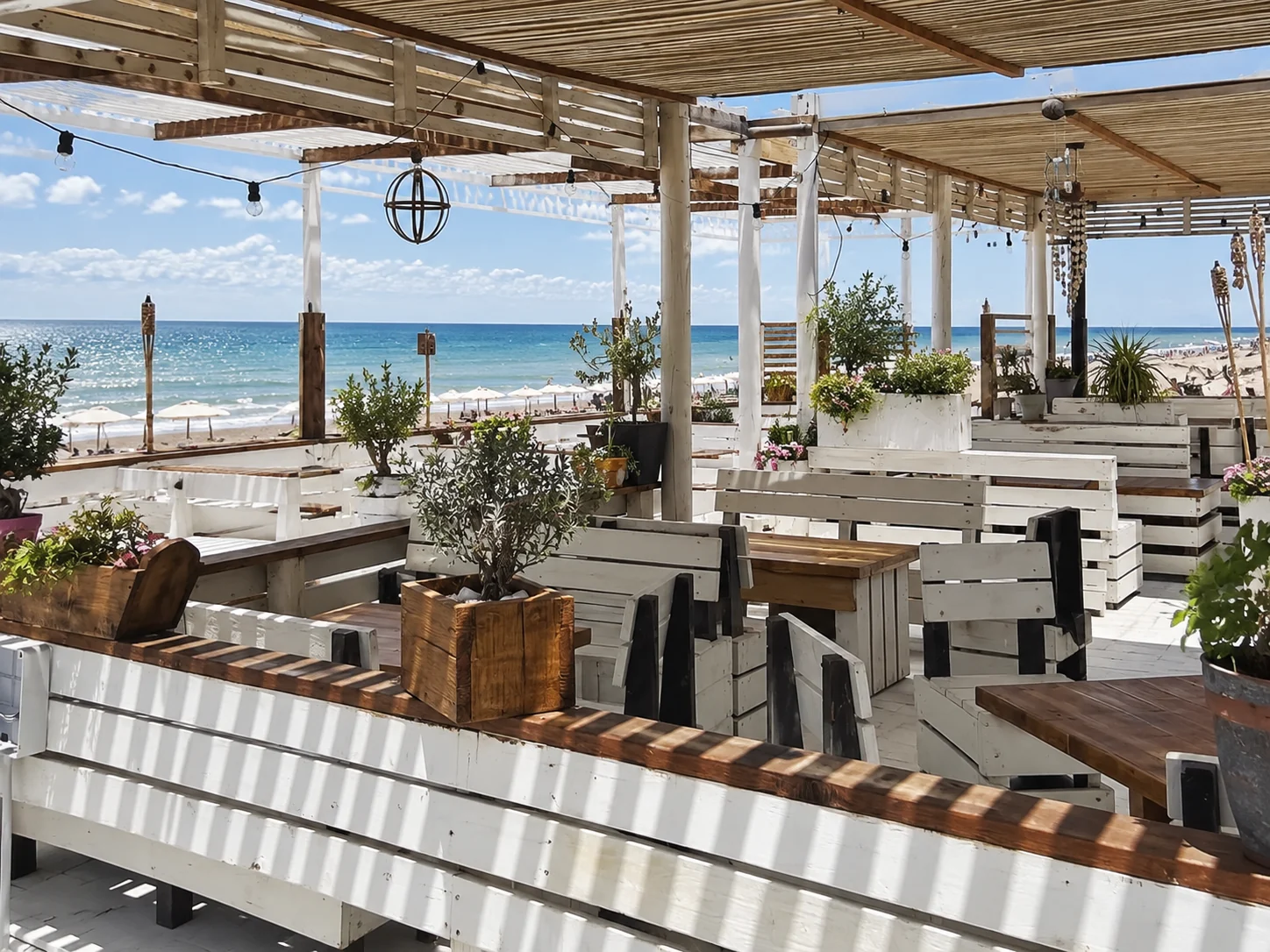 Visualización de la terraza de SHANTI: pérgola, mesas y bancos blancos frente al mar y al horizonte.